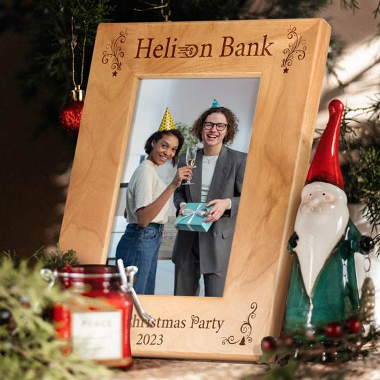 A personalized red alder wood frame engraved with logo for ‘Helixon Bank’ and ‘Christmas Party 2023’ engraved at the bottom, displayed on a tabletop with holiday decorations including a gnome figurine, a red candle jar, pine branches, and ornaments. The frame contains a picture of 2 co-workers celebrating at a party.