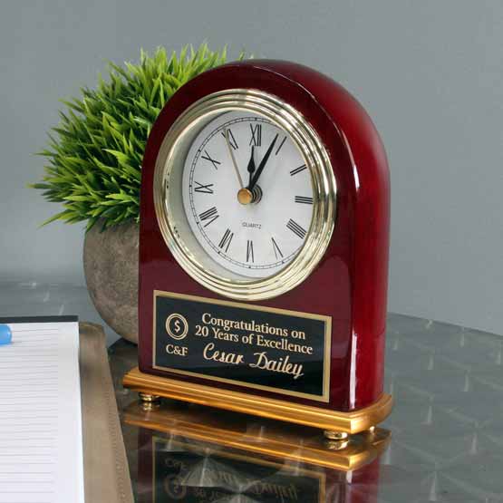 Rosewood Portrait Desk Clock With Gold Trim And Metal Plate With Years Of Excellence Message Standing On Table By Plant