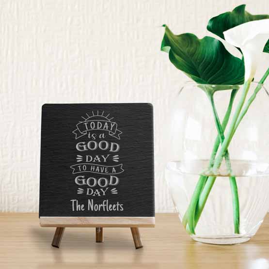 Custom Slate Sign Printed With Good Day Today Message And Family Name Standing Via Wood Easel On Shelf With Plant In Vase