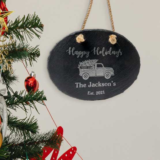Slate Oval Sign Printed With Happy Holidays And Name And Date And Festive Truck Artwork Hanging By Christmas Tree Via Rope
