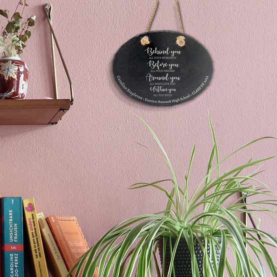 Slate Oval Sign Printed With Inspiring Message And Student Graduation Details Hanging Via Rope By Shelf And Plants And Books