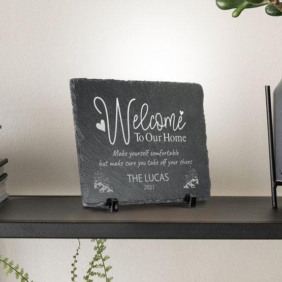 A personalized dark gray slate stone sign on a black easel stand, displayed on a shelf. The sign features engraved light gray text reading ‘Welcome To Our Home’ and “THE LUCAS 2021’.