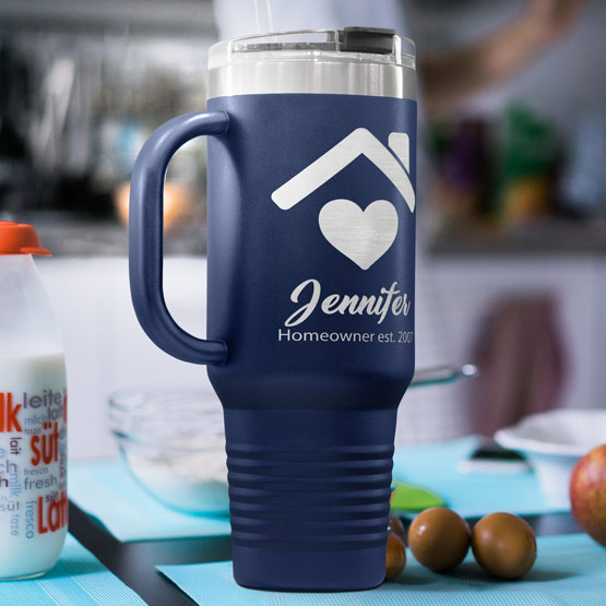 A navy blue insulated handle tumbler mug with a clear lid, featuring engraved house and heart graphic and the personalized text ‘Jennifer Homeowner est. 2021’, set on a kitchen counter with a blurred background.