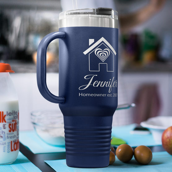 A navy blue insulated handle tumbler mug with a clear lid, featuring engraved house and heart graphic and the personalized text ‘Jennifer Homeowner est. 2021’, set on a kitchen counter with a blurred background.