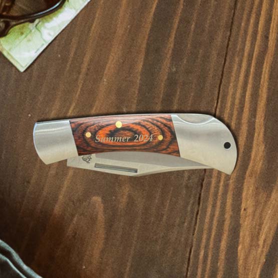 Custom Stainless Steel Folding Knife With Wood Style Handle And Engraved With Summer 2024 On A Wood Table Near Sunglasses