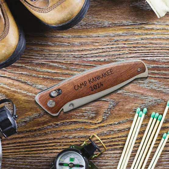 Custom Wood And Stainless Steel Folding Knife With Button Lock And Engraved With Camp Name On Wood Table With Outdoorsy Tools