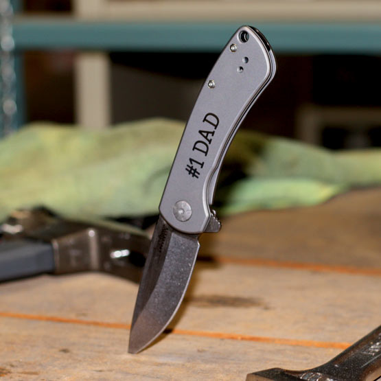 Custom Stainless Steel Folding Knife Laser Fused With Number One Dad Message Piercing A Wood Work Table Near Tools
