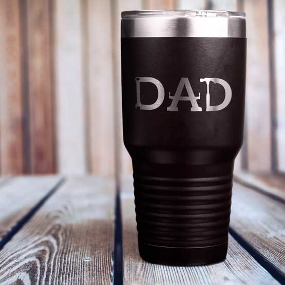 Custom Black Thirty Ounce Coffee Tumbler With Straw And Engraved With Tools Art Spelling Out Dad Text On A Wood Table