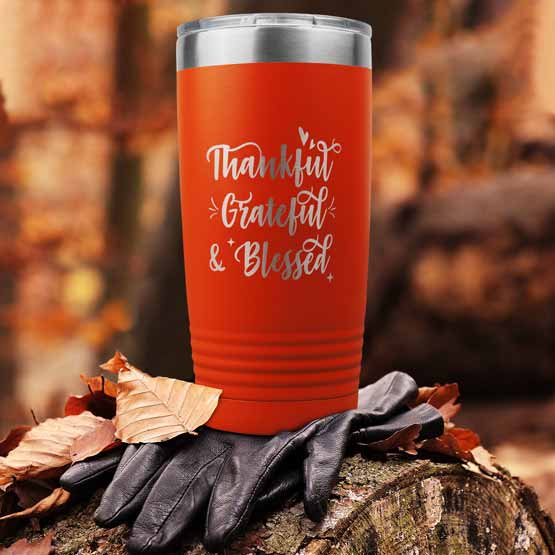 Orange 20 Ounce Tumbler Engraved With Cursive Thankful Grateful And Blessed Message Sitting Outdoors On Gloves And Forest Log