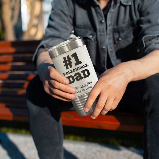 Man On Bench Holding Faux Leather Wrapped Coffee Tumbler With Volleyball Theme And Engraved With Number One Dad Message
