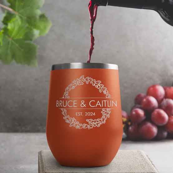 Wine Bottle Pouring Wine Into Orange Stemless Wine Tumbler Engraved With Couple Names And Wheat Art On A Kitchen Counter