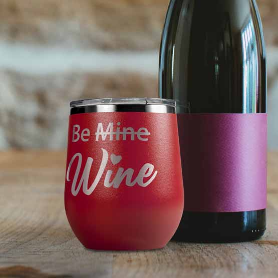 Red 12 Ounce Stemless Wine Tumbler Engraved With Be Mine Crossed Out Above Cursive Wine Text On Table Beside Wine Bottle