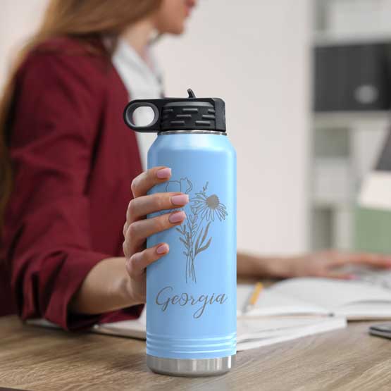 Woman At Office Desk Holding Light Blue Thirty Two Ounce Water Bottle With Straw And Laser Engraved With Name And Flower Art