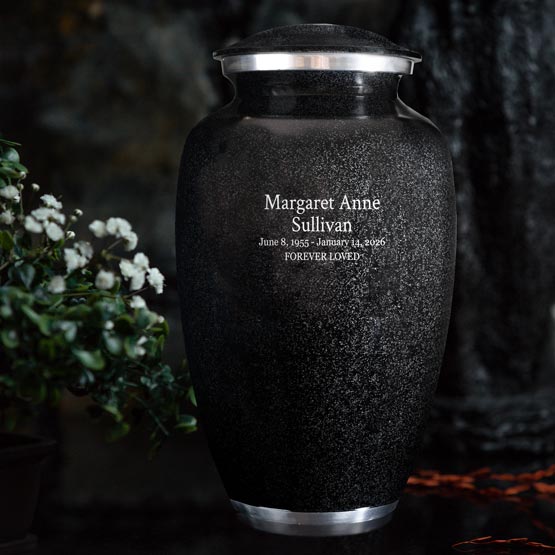 Black Aluminum Cremation Urn Engraved With Custom Memorial Details And Message Sitting On A Table Beside White Flowers