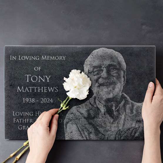 Woman Hands Indoors Placing Flowers Onto Rectangle Granite Headstone Engraved With Image Of Older Man And Memorial Details