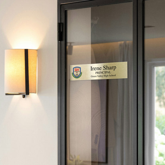 A gold metal office plate displayed on a glass door indicating the office is the principal’s office of Irene Sharp. A rectangular wall sconce with warm light, mounted on wall next to the glass door