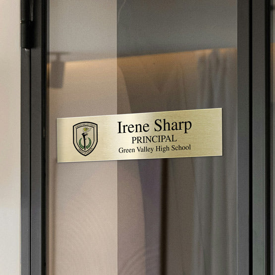 A gold metal office plate displayed on a glass door indicating the office is the principal’s office of Irene Sharp. A rectangular wall sconce with warm light, mounted on wall next to the glass door