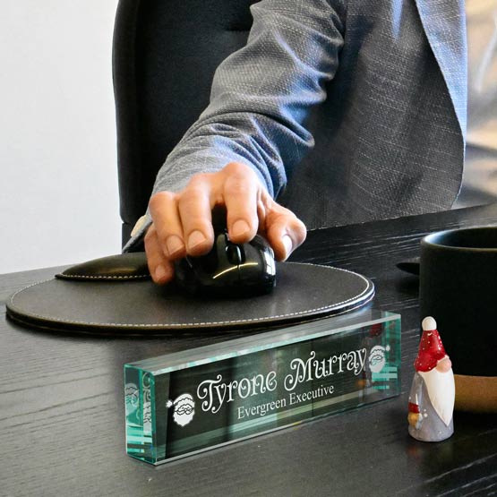 A glass nameplate displaying ‘Tyrone Murray Evergreen Executive’ sits on a dark wooden desk next to a small Santa figurine. A person in a gray blazer, sits in the background using the computer.