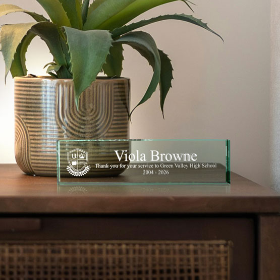 A jade tinted glass desk plate engraved with white text that reads ‘Viola Browne’ and a school crest, placed on a wooden cabinet next to a potted plant in a beige ceramic pot.