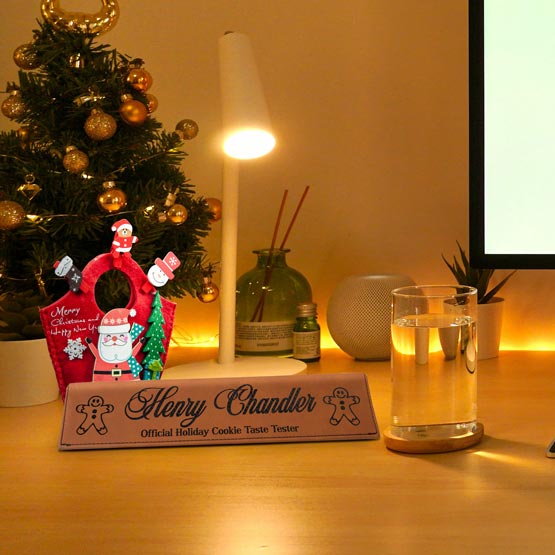 A personalized brown faux leather desk wedge with engraved text that reads ‘Henry Chandler Official Holiday Cooke Taste Tester’. The desk wedge is displayed on a wooden desk with a Santa decoration and Christmas tree in the background, under a warm light desk lamp.