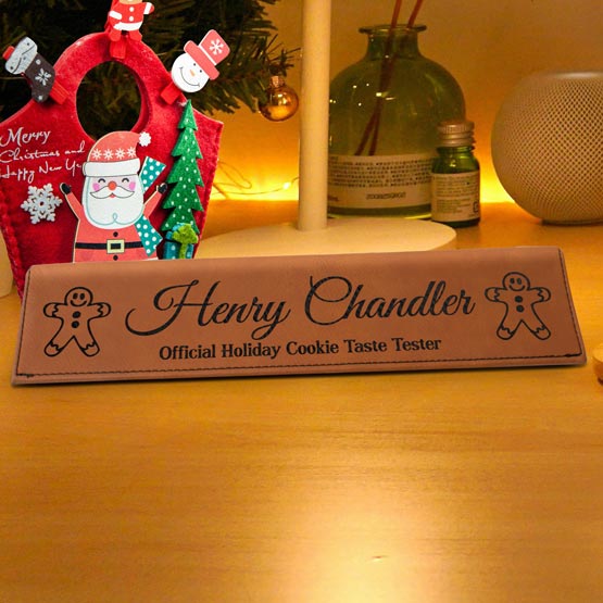 A personalized brown faux leather desk wedge with engraved text that reads about an official Holiday cookie taste tester. The desk wedge is displayed on a wooden desk with a Santa decoration and Christmas tree in the background.