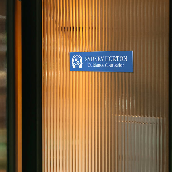 A sky blue plastic name plate read ‘Sydney Horton Guidance Counselor’ with a white head icon, mounted on a warm amber ribbed glass door.