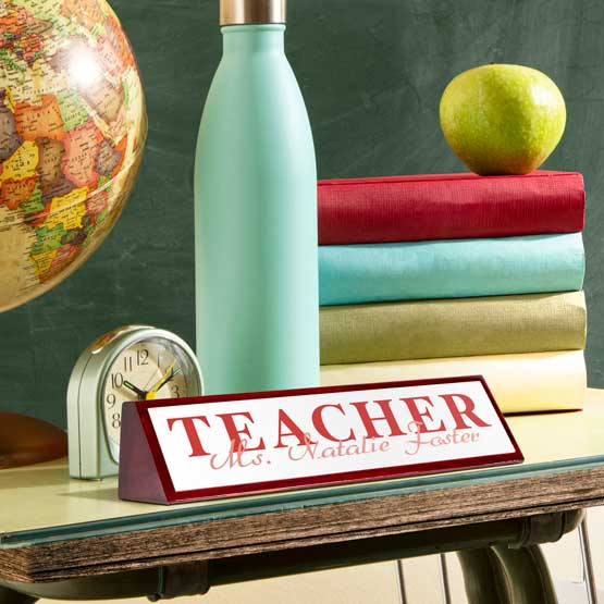 Desk setup with 'TEACHER' nameplate, books, apple, globe, and water bottle on a chalkboard background.