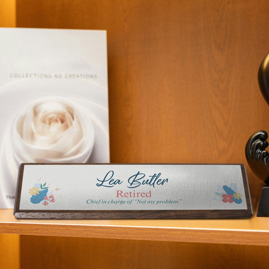 A personalized walnut wooden desk wedge with a silver metal plate mounted on the front, reading ‘Lea Butler Retired’ with a humorous quote, displayed on a wooden shelf.