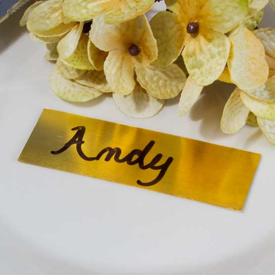 Brass Name Tag From Set Of Ten Marked With Handwritten Andy Text In Black On A White Table With Yellow Flowers