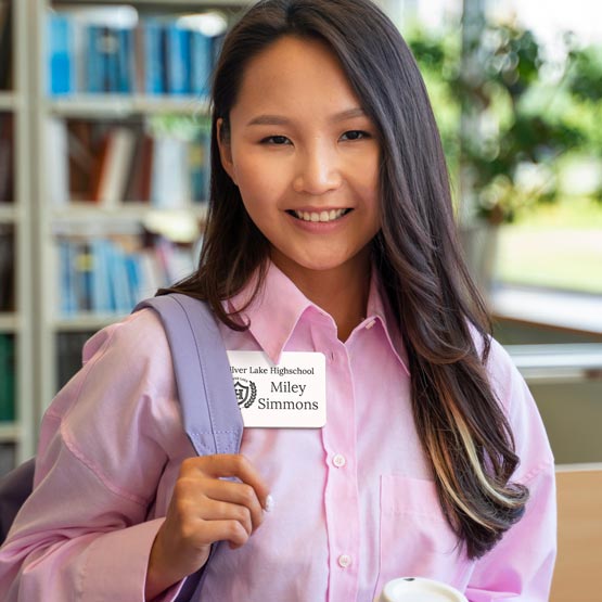A smiling young woman with long dark hair stands in a library or study, wearing pink button up shirt with a lavender shoulder bag. She has a white plastic name tag that reads ‘Silver Lake High School Miley Simmons’. 