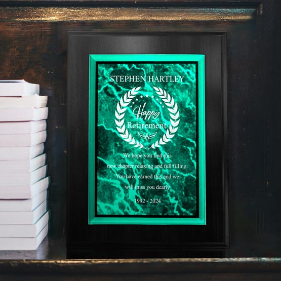 A personalized retirement plaque with green acrylic plate with a marble texture on the front mounted to a black wooden board, white engraved text reads ‘Stephen Hartley’ and ‘Happy Retirement’. The plaque is displayed on a dark wooden surface next to a stack of white books.