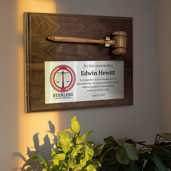 A personalized plaque featuring a gavel mounted on a walnut wood board, with a silver metal plate printed with a Sterling Justice Martini logo and text. The plaque is mounted on a beige wall above green plants, with warm sunlight casting a shadow on the left.