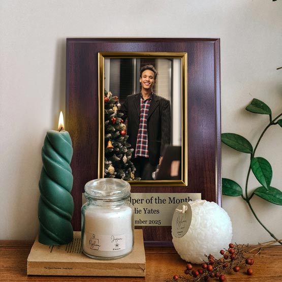 A personalized cherry wood board picture frame with gold frame and gold metal tag at the bottom with text, displaying a smiling man wearing a Santa hat. The frame is on a wooden surface next to a green twist candle, a white jar candle, a white fluffy ornament, and a small pant against a neutral wall