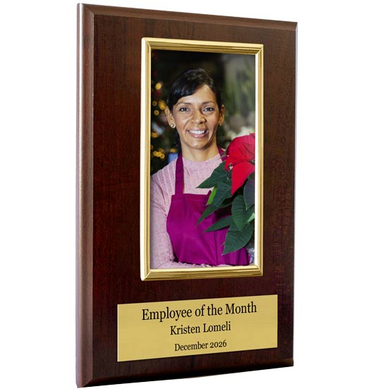 A vertical wooden plaque with a dark cherry wood finish, featuring a gold-framed photo of a smiling woman in a purple apron holding red poinsettias, and a gold nameplate at the bottom reading 'Employee of the Month, Kristen Romeli, December 2026'.