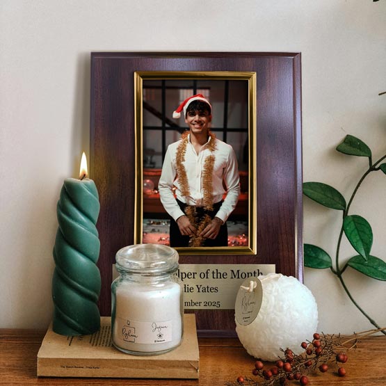 A personalized cherry wood board picture frame with gold frame and gold metal tag at the bottom with text, displaying a smiling man wearing a Santa hat. The frame is on a wooden surface next to a green twist candle, a white jar candle, a white fluffy ornament, and a small pant against a neutral wall