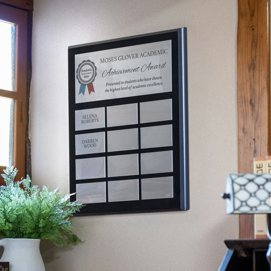 A coated black board perpetual plaque with silver plates with an academic achievement award mounted on a beige wall in an office setting. Featuring a personalized designed printed on the top plate and multiple blank plates below, two plates have names printed.