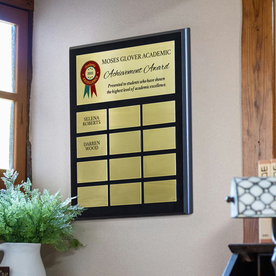 A coated black board perpetual plaque with gold plates with an academic achievement award mounted on a beige wall in an office setting. Featuring a personalized designed printed on the top plate and multiple blank plates below, two plates have names printed.