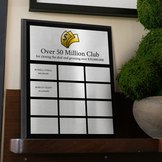 A custom corporate perpetual plaque with silver aluminum metal plates. The top plate features black text which reads ‘Over 50 Million Club’ with a colorful logo above. The smaller plates below it have two plates filled in with names and the rest are blank displayed on a wooden shelf next to a green potted plant.