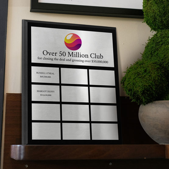A custom corporate perpetual plaque with silver aluminum metal plates. The top plate features black text which reads ‘Over 50 Million Club’ with a colorful logo above. The smaller plates below it have two plates filled in with names and the rest are blank displayed on a wooden shelf next to a green potted plant.