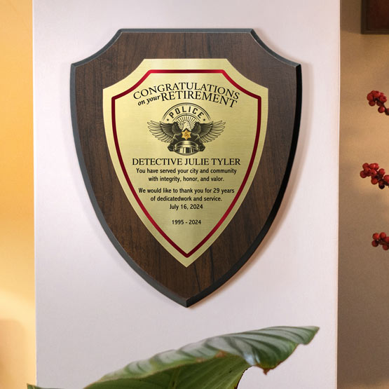 A personalized wooden shield-shaped wall plaque for a retired police detective, mounted on a beige wall. The plaque has a gold metal plate attached to a cherry wood board with full color printed text.
