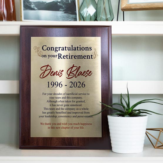 A gold metal retirement commemorative plaque mounted on a coated cherry board, displayed on a shelf next to a small potted plant. The plaque is printed in full color with text congratulating Denis Blase on retirement.
