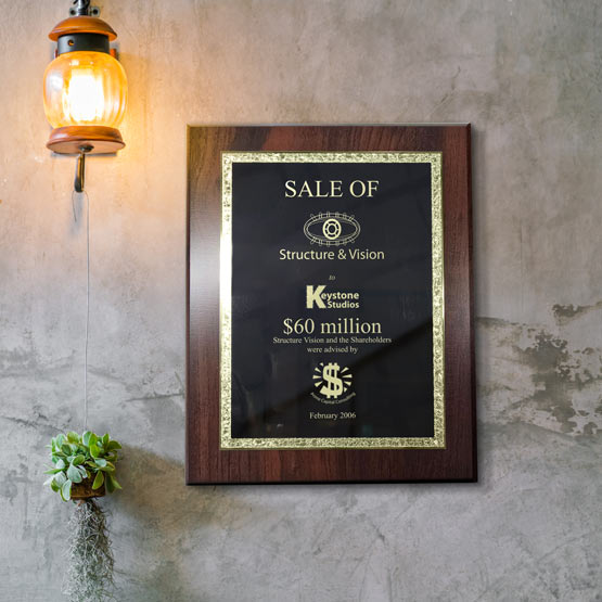 A black metal award of sales plaque with a decorative gold trim board and engraved text, mounted on a textured gray wall under a hanging amber lantern light.