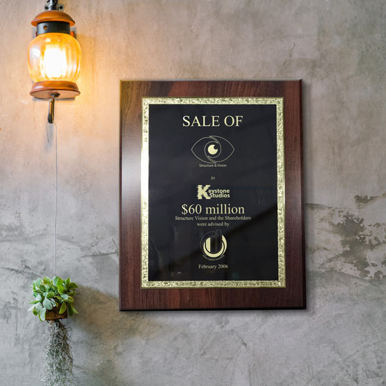 A black metal award of sales plaque with a decorative gold trim board and engraved text, mounted on a textured gray wall under a hanging amber lantern light.