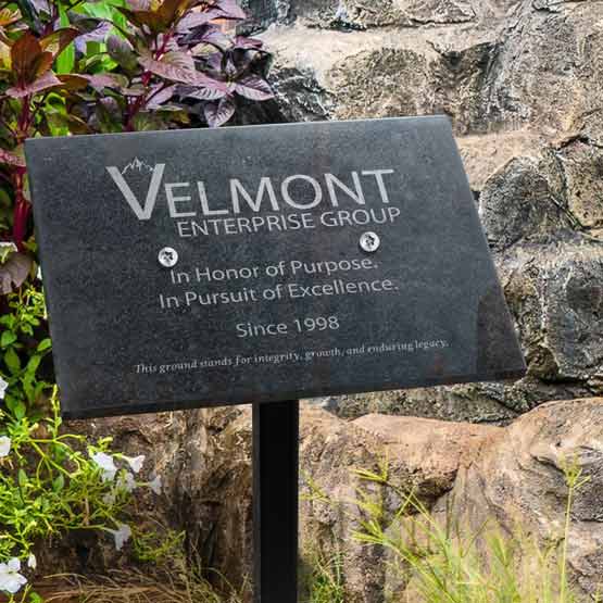 Granite Plaque Engraved With Custom Company Logo And Details Displayed Via Sign Stand Outdoors Near Rocks And Plants
