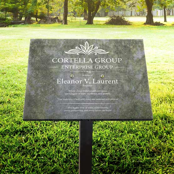 A granite plaque attached to a metal stand, displayed in grass with trees and a park in the background. The plaque features engraved light gray text honoring Eleanor V. Laurent.