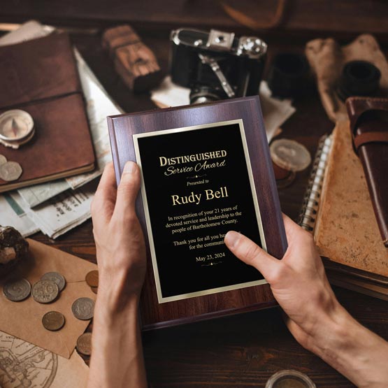 A person holds a personalized laser metal plaque with a coated cherry board reading 'Distinguished Service Award' and 'Rudy Bell.' The scene is a styled desk setup with a vintage camera, notebooks, coins, and papers around it.