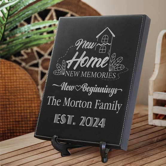 A personalized black faux leather plaque on a small black easel stand, displayed on a wooden table in a home setting. The sign features silver engraved text reading ‘New Home NEW MEMORIES’, ’New Beginnings’, ‘The Morton Family’, and ‘EST 2024’.