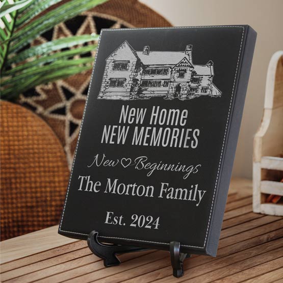 A personalized black faux leather plaque on a small black easel stand, displayed on a wooden table in a home setting. The sign features silver engraved text reading ‘New Home NEW MEMORIES’, ’New Beginnings’, ‘The Morton Family’, and ‘EST 2024’.