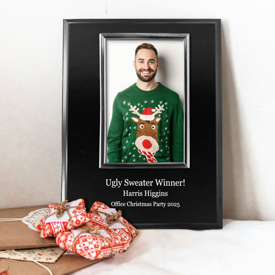 A personalized photo frame on a black board with silver frame next to wrapped Christmas gifts. The frame contains a picture of a smiling man in a green Christmas sweater with a reindeer graphic, with text printed on the board that reads ‘Ugly Sweater Winner! Harris Higgins Office Christmas Party 2025’.
