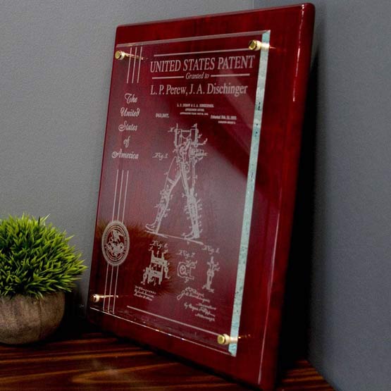 Floating Glass Plaque With A Rosewood Board And Frosted Engravings Of Patent Details On Shelf With Plant Leaning Against Wall